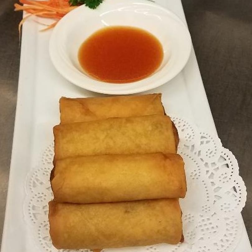 Four golden-brown Thai spring rolls are stacked on a white rectangular plate with a side of dipping sauce.