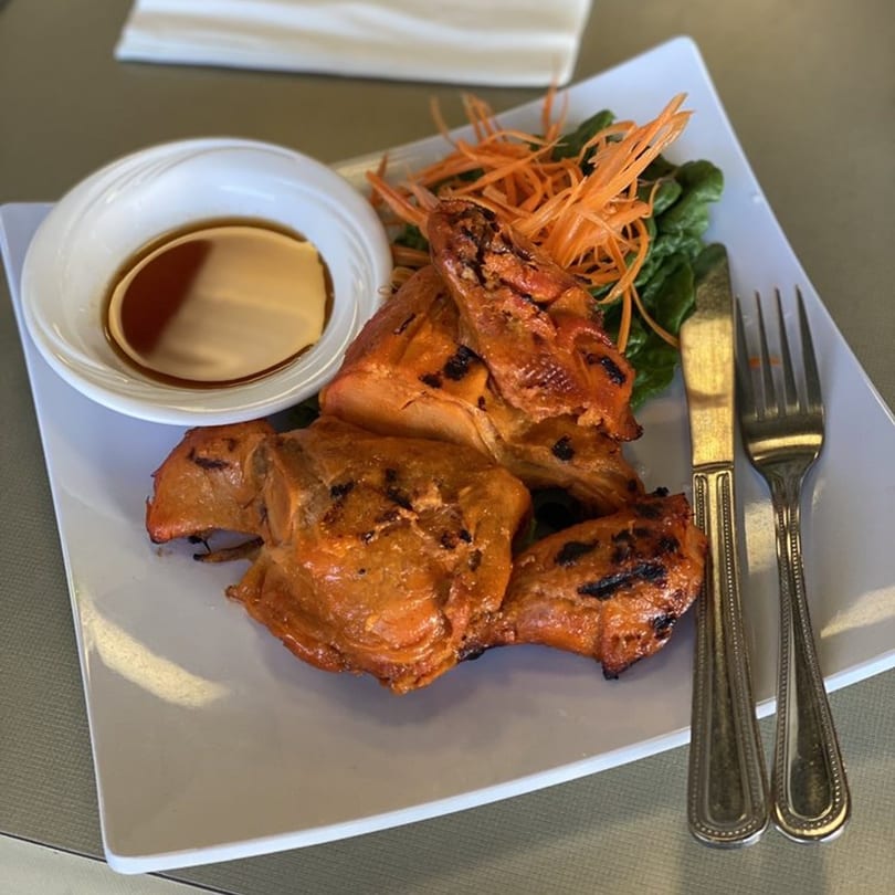 Thai BBQ chicken with shredded carrots, greens, and dipping sauce on a white plate.